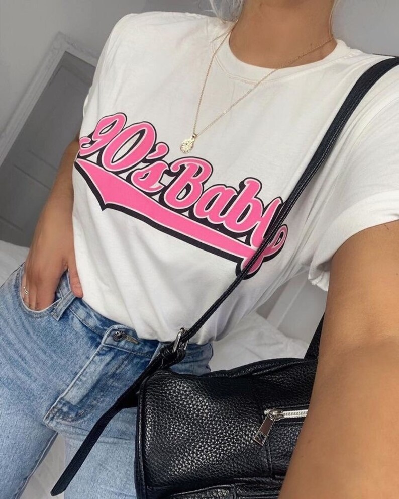 May include: White t-shirt with a pink "90's Baby" graphic. The shirt is worn with blue jeans and a black leather crossbody bag.