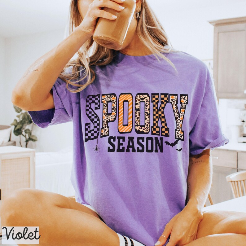 Może przedstawiać: Fioletowa koszulka z napisem "SPOOKY SEASON" w połączeniu z kratą, panterką i pomarańczowymi literami. Mały pająk i grafika nietoperza znajdują się pod tekstem. Słowo "Violet" znajduje się w lewym dolnym rogu.