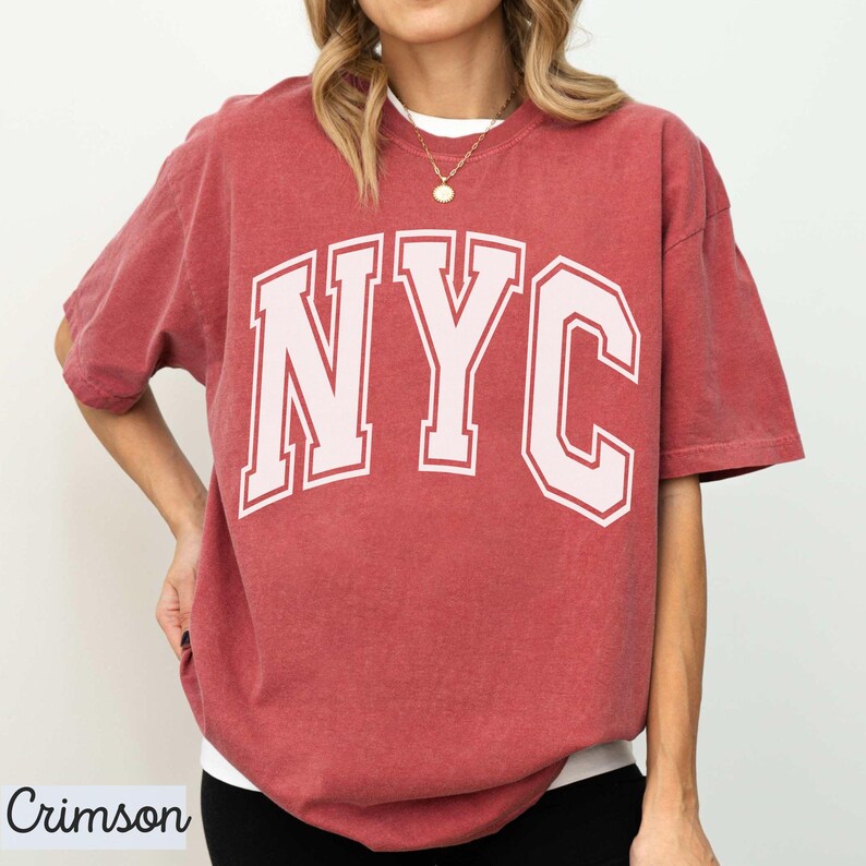 May include: Crimson-colored t-shirt with the white letters "NYC" in a collegiate font. The shirt has short sleeves and a relaxed fit. The word "Crimson" is printed in a script font in the lower left corner.