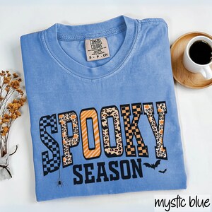 Może przedstawiać: Jasnoniebieska koszulka z napisem "SPOOKY SEASON" w halloweenowym wzorze. Słowo "SPOOKY" ma wz&oacute;r w szachownicę, nadruk w cętki i pomarańczowe paski. W projekcie uwzględniono r&oacute;wnież pająka i nietoperze.