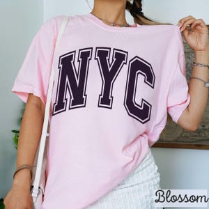 May include: A light pink t-shirt with the letters "NYC" in a dark purple collegiate font. The shirt is short-sleeved and appears to be made of a soft material. The word "Blossom" is printed in white at the bottom right corner.