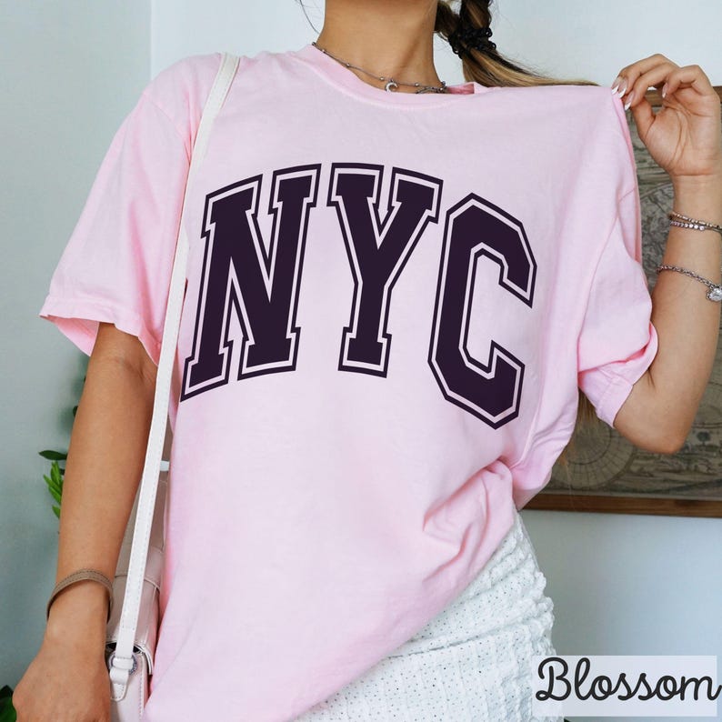 May include: A light pink t-shirt with the letters "NYC" in a dark purple collegiate font. The shirt is short-sleeved and appears to be made of a soft material. The word "Blossom" is printed in white at the bottom right corner.
