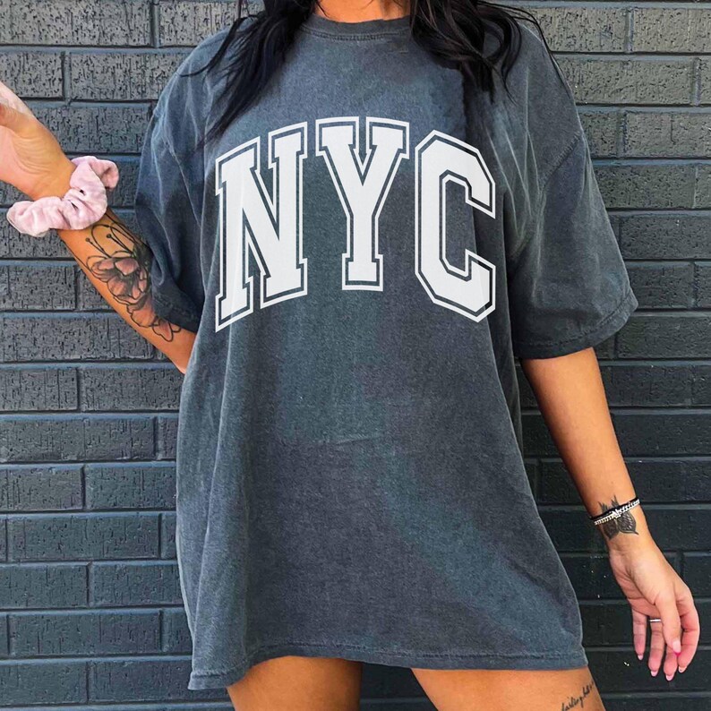 May include: A charcoal gray oversized t-shirt with the white letters "NYC" in a collegiate font. The shirt has short sleeves and a slightly faded appearance. The image is set against a dark gray brick wall.