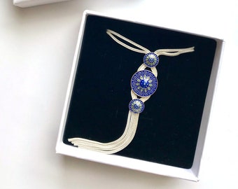May include: A silver-toned necklace with three circular blue and silver decorative elements, presented in a white box with the word "Mesopotamia" printed on it. The necklace has a tassel-like design.