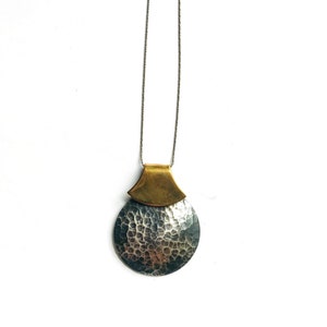May include: A silver-toned hammered metal pendant necklace with a brass-colored top piece. The pendant is circular with a textured surface, suspended from a delicate silver chain. The necklace is displayed against a white background, highlighting the contrast of the metals.