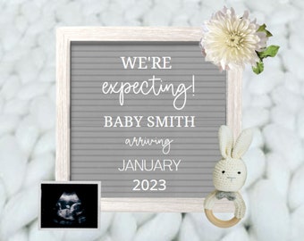 May include: A gray letter board with white letters announcing "We're expecting! Baby Smith arriving January 2023". A white crocheted bunny with a wooden ring is on the right side of the board. A white flower is on the top right corner of the board. A sonogram image is on the bottom left corner of the board.
