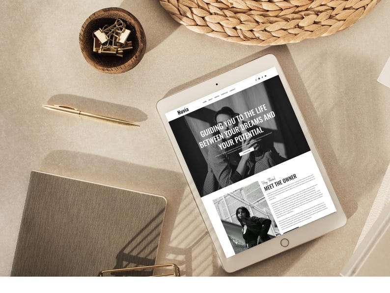 May include: A tablet displaying a website with the text "Guiding you to the life between your dreams and your potential." A gold pen, a small wooden bowl with gold paper clips, and a notebook are also on the beige surface.