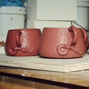 May include: Three terracotta-coloured ceramic mugs with a textured, dimpled surface. Each mug features a handle and a decorative, coiled snake-like clay element attached to the lower side. The mugs are arranged in a row on a flat wooden board.