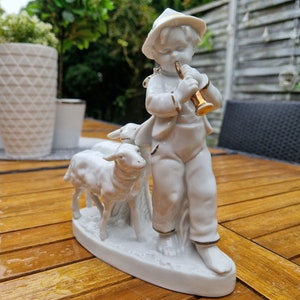 May include: A white porcelain figurine of a boy playing a flute, standing with two white porcelain lambs. The boy is wearing a hat and has a gold trim on his clothing.