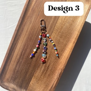 Pu&ograve; includere: Un portachiavi di perline con chiusura color oro. Il portachiavi presenta perline colorate di varie forme e dimensioni, comprese perline trasparenti a forma di cuore. Il testo "Design 3" &egrave; in un rettangolo bianco.