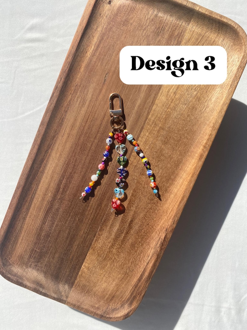 Pu&ograve; includere: Un portachiavi di perline con chiusura color oro. Il portachiavi presenta perline colorate di varie forme e dimensioni, comprese perline trasparenti a forma di cuore. Il testo "Design 3" &egrave; in un rettangolo bianco.