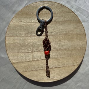 May include: A brown and red beaded keychain with a silver key ring. The keychain is made of macrame and has a small red bead with a white design.