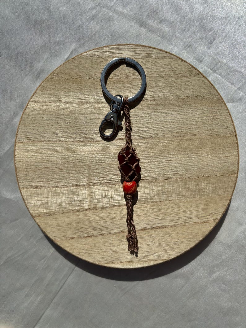 May include: A brown and red beaded keychain with a silver key ring. The keychain is made of macrame and has a small red bead with a white design.