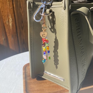 Pu&ograve; includere: Borsa verde oliva con portachiavi di perline. Il portachiavi presenta perline colorate, un ciondolo a forma di cuore dorato con la lettera "C" e un fermaglio argentato. La borsa ha una barra argentata e il nome del marchio in rilievo sul lato.