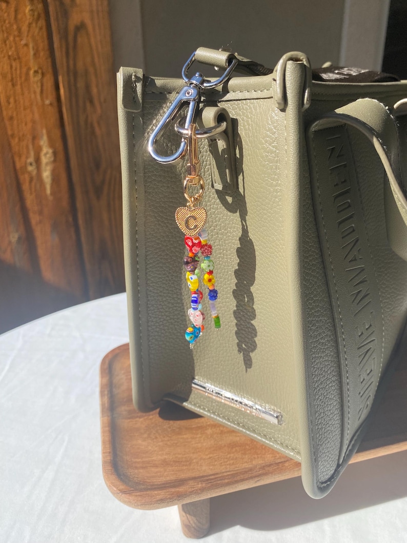 Pu&ograve; includere: Borsa verde oliva con portachiavi di perline. Il portachiavi presenta perline colorate, un ciondolo a forma di cuore dorato con la lettera "C" e un fermaglio argentato. La borsa ha una barra argentata e il nome del marchio in rilievo sul lato.