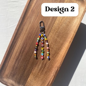 Pu&ograve; includere: Un portachiavi di perline con chiusura argentata. Il portachiavi presenta fili di perline colorate, tra cui rosso, giallo, blu e verde, con disegni floreali e fantasia. Il testo "Design 2" &egrave; in un rettangolo bianco.