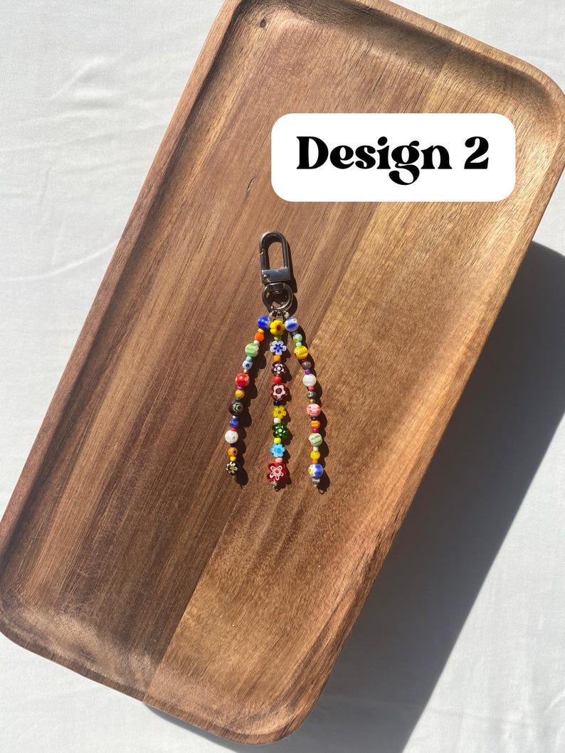 Pu&ograve; includere: Un portachiavi di perline con chiusura argentata. Il portachiavi presenta fili di perline colorate, tra cui rosso, giallo, blu e verde, con disegni floreali e fantasia. Il testo "Design 2" &egrave; in un rettangolo bianco.