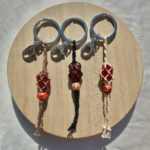 May include: Three keychains with red and orange gemstone accents. Each keychain has a silver ring and a macrame tassel. The tassels are brown, black, and white.