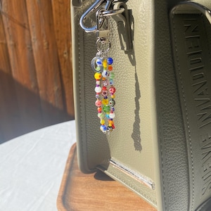 Pu&ograve; includere: Un portachiavi di perline colorate con chiusura argentata &egrave; appeso a una borsa in pelle verde. Il portachiavi presenta una variet&agrave; di piccole perline rotonde nei toni del rosso, giallo, blu e verde. La borsa ha una barra argentata e le parole "STEVE MADDEN" in rilievo sul lato.