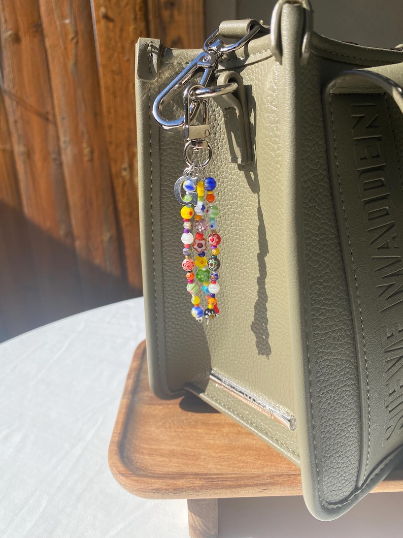 Pu&ograve; includere: Un portachiavi di perline colorate con chiusura argentata &egrave; appeso a una borsa in pelle verde. Il portachiavi presenta una variet&agrave; di piccole perline rotonde nei toni del rosso, giallo, blu e verde. La borsa ha una barra argentata e le parole "STEVE MADDEN" in rilievo sul lato.