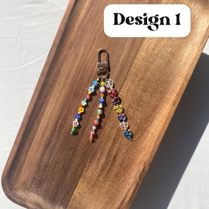 Pu&ograve; includere: Un portachiavi di perline con chiusura dorata. Il portachiavi presenta tre fili di perline colorate, tra cui forme di fiori e cuori. Il testo "Design 1" &egrave; in un rettangolo bianco in alto a destra. Il portachiavi &egrave; appoggiato su un vassoio di legno.