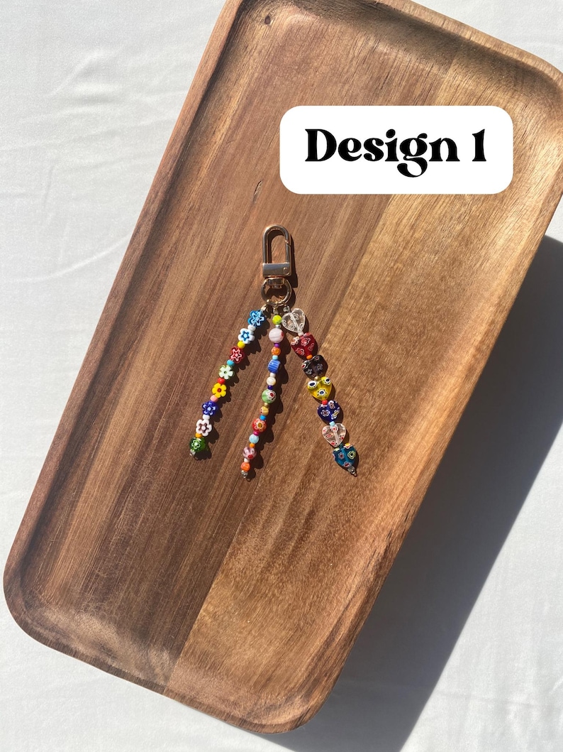 Pu&ograve; includere: Un portachiavi di perline con chiusura dorata. Il portachiavi presenta tre fili di perline colorate, tra cui forme di fiori e cuori. Il testo "Design 1" &egrave; in un rettangolo bianco in alto a destra. Il portachiavi &egrave; appoggiato su un vassoio di legno.