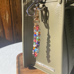 Pu&ograve; includere: Un portachiavi di perline colorate con chiusura dorata &egrave; appeso a una borsa verde. Il portachiavi presenta piccole perline rotonde rosse, blu, arancioni e gialle. La borsa verde oliva ha una barra argentata sul davanti.