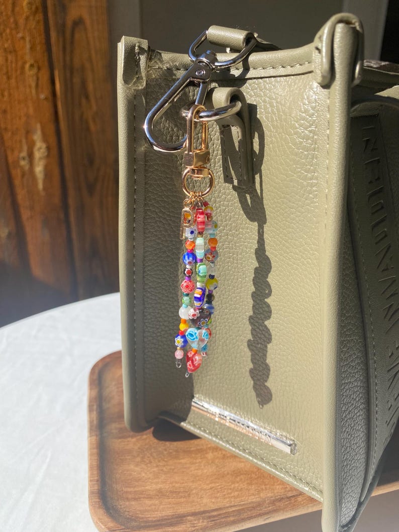Pu&ograve; includere: Un portachiavi di perline colorate con chiusura dorata &egrave; appeso a una borsa verde. Il portachiavi presenta piccole perline rotonde rosse, blu, arancioni e gialle. La borsa verde oliva ha una barra argentata sul davanti.