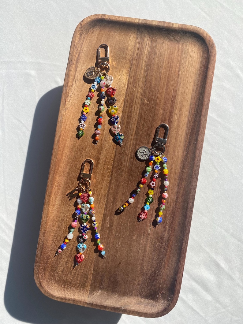 Pu&ograve; includere: Tre portachiavi con perline, chiusure color oro e piccoli ciondoli. I portachiavi presentano perline colorate di varie forme, tra cui fiori e cuori. Sono disposti su un vassoio di legno.