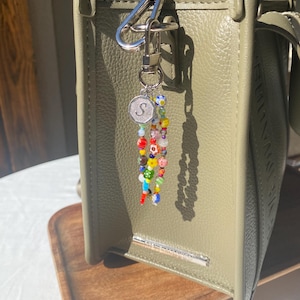 Pu&ograve; includere: Una borsa in pelle verde con chiusura in metallo argentato e un portachiavi con perline. Il portachiavi presenta perline colorate e un ciondolo argentato con la lettera "S". La borsa &egrave; appoggiata su un vassoio di legno.