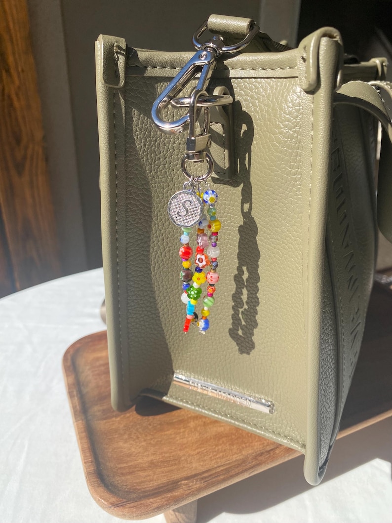 Pu&ograve; includere: Una borsa in pelle verde con chiusura in metallo argentato e un portachiavi con perline. Il portachiavi presenta perline colorate e un ciondolo argentato con la lettera "S". La borsa &egrave; appoggiata su un vassoio di legno.