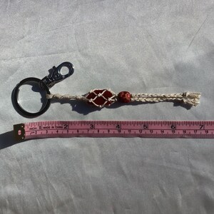 May include: A red and white macrame keychain with a red stone bead and a silver key ring. The keychain is about 6 inches long.
