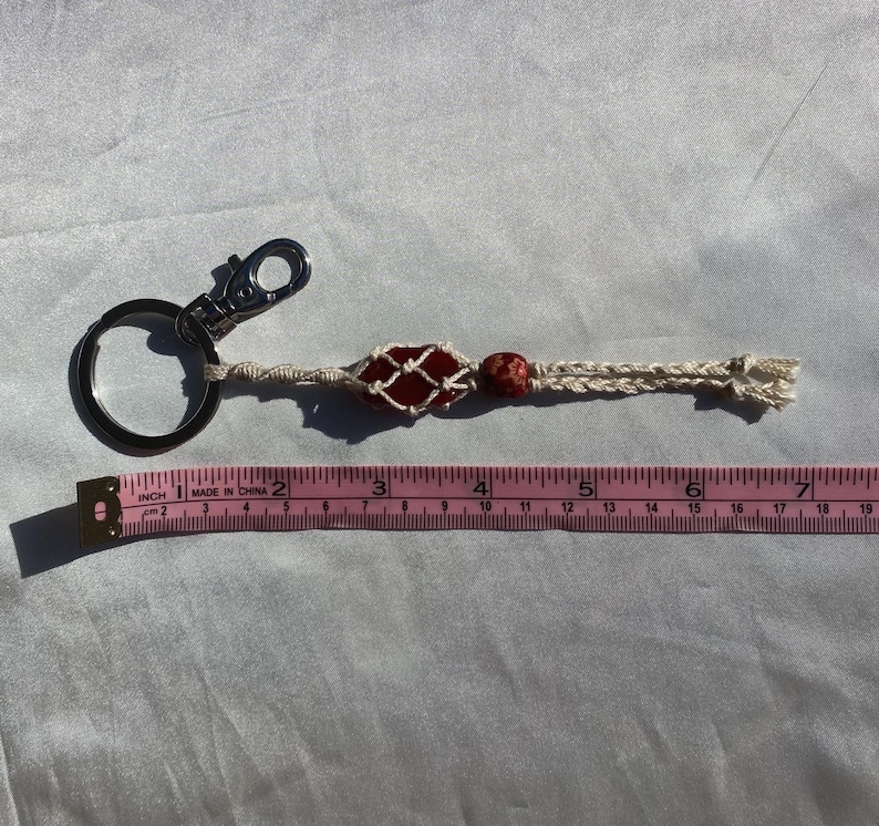 May include: A red and white macrame keychain with a red stone bead and a silver key ring. The keychain is about 6 inches long.
