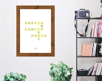 May include: A framed print with a white background and a yellow heart in the bottom centre. The print features the names Amelia, Family, and David spelled out using Scrabble tiles.