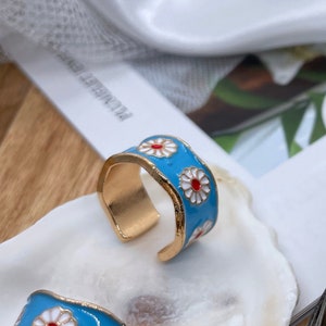 May include: Four gold ear cuffs with a blue enamel base and white daisy designs. The cuffs are displayed on a white and brown seashell.