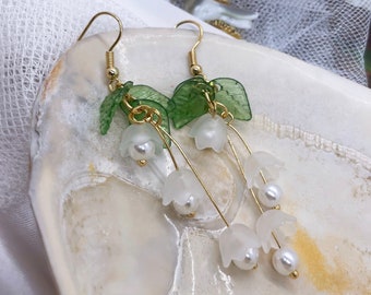 May include: A pair of gold earrings with white flower charms and green leaves. The earrings are hanging from a white seashell.