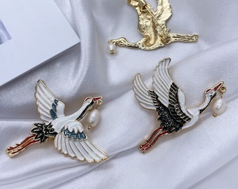 Puede incluir: Tres broches de grulla de tono dorado con detalles de perla. Las grullas están en vuelo y tienen detalles de esmalte negro, blanco, rojo y azul.