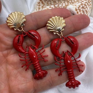 May include: A pair of red lobster earrings with gold-colored seashell studs. The earrings are a bright red color and feature detailed lobster designs. The earrings are a fun and unique accessory.