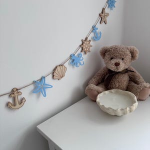 May include: A blue and brown nautical themed garland with anchors, seashells, starfish, and steering wheels hanging on a white wall. A brown teddy bear sits on a white table with a white candle in a shell-shaped holder.