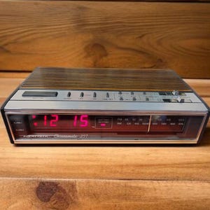 May include: A vintage Realistic Chronomatic 257 digital alarm clock. The clock has a brown and silver top with a red LED display showing the time as 12:15. It features AM/FM radio and alarm functions. The clock is set on a wooden surface.