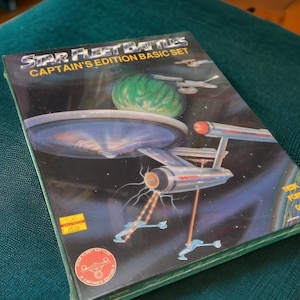 Puede incluir: Una caja de juego de mesa Star Fleet Battles Captain's Edition Basic Set con una ilustraci&oacute;n de una nave espacial y un planeta. La caja es azul, negra y blanca con el texto "Star Fleet Battles Captain's Edition Basic Set" y "The Original Star Fleet Battles Game".