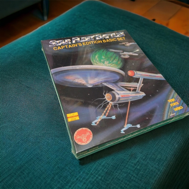 Puede incluir: Una caja de juego de mesa Star Fleet Battles Captain's Edition Basic Set con una ilustraci&oacute;n de una nave espacial y un planeta. La caja es azul, negra y blanca con el texto "Star Fleet Battles Captain's Edition Basic Set" y "The Original Star Fleet Battles Game".