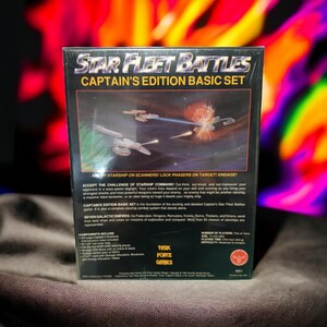 Puede incluir: Una caja de juego de mesa Star Fleet Battles Captain's Edition Basic Set con un fondo negro y una imagen colorida de dos naves espaciales en una batalla. La caja presenta el texto "Star Fleet Battles" y "Captain's Edition Basic Set".