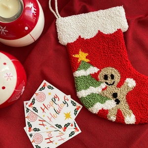 May include: A red and white Christmas stocking with a gingerbread man and a green Christmas tree. The stocking is decorated with white fluffy trim and is sitting on a red fabric surface. There are three cards that say "Happy Holidays" in pink and white lettering.