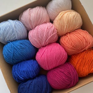 May include: A cardboard box filled with colorful yarn balls. The yarn is in various shades of pink, blue, orange, and white. The yarn balls are neatly arranged, showcasing a variety of colors suitable for knitting or crocheting projects.