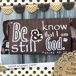 May include: A brown paper wrapper for a candy package featuring the white printed text Be still & know that I am God and Psalms 46:10. The wrapper has a circular cutout in the center revealing a dark brown candy underneath the paper cover.