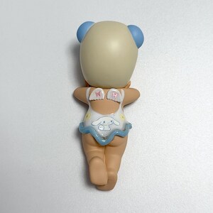 May include: A figurine of a character with a light tan head and blue ear-like features. The figurine is wearing a white swimsuit with a blue frill and a cartoon character design. The figurine is posed with its arms outstretched.