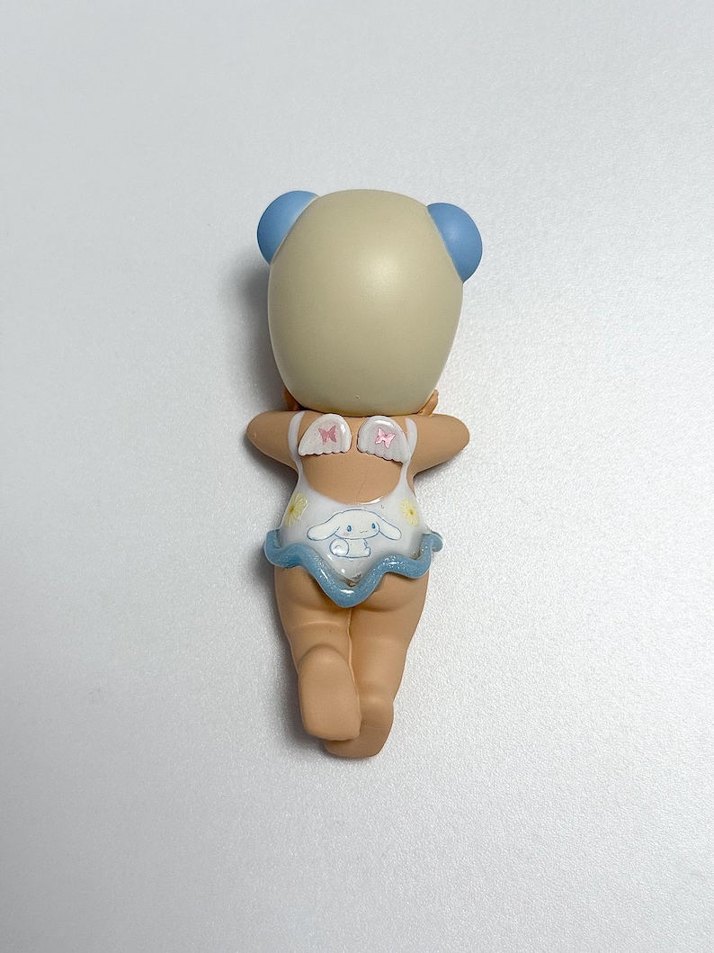 May include: A figurine of a character with a light tan head and blue ear-like features. The figurine is wearing a white swimsuit with a blue frill and a cartoon character design. The figurine is posed with its arms outstretched.