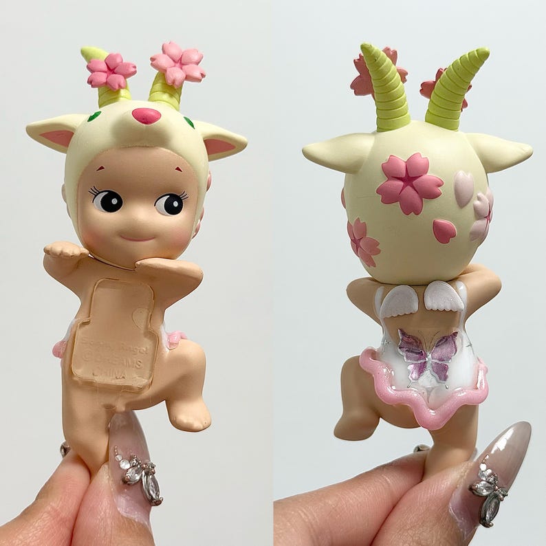 May include: A small, cream-coloured figurine of a baby goat wearing a hood with pink flower accents. The figurine has a pink tutu and a butterfly design on the back. The goat has green horns and pink flowers on its head.