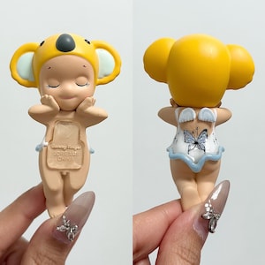 May include: A figurine of a child wearing a yellow koala hat and a light-coloured outfit. The figurine has closed eyes and is holding its hands up to its face. The back of the figurine has a butterfly design on the outfit.